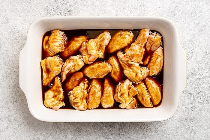 A white rectangular baking dish filled with pieces of raw chicken coated in a dark brown marinade, placed on a light gray textured surface.