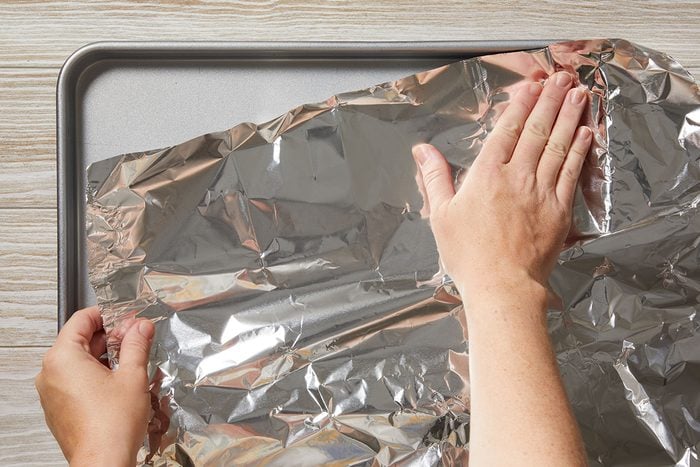 Two hands are spreading a sheet of aluminum foil over a rectangular baking tray on a light-colored wooden surface.