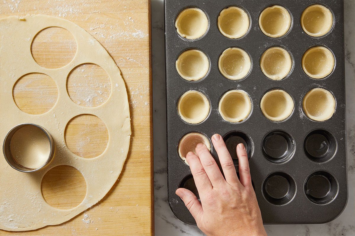 Overhead shot of round dough cutouts placed in a muffin tin, hand pressing one piece into the mold.