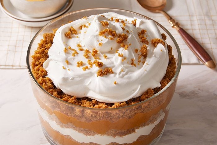 A layered dessert in a glass bowl, featuring alternating layers of whipped cream and a brown crumb mixture, topped with whipped cream and sprinkled crumbs. A spoon and a bowl are in the background on a marble surface.
