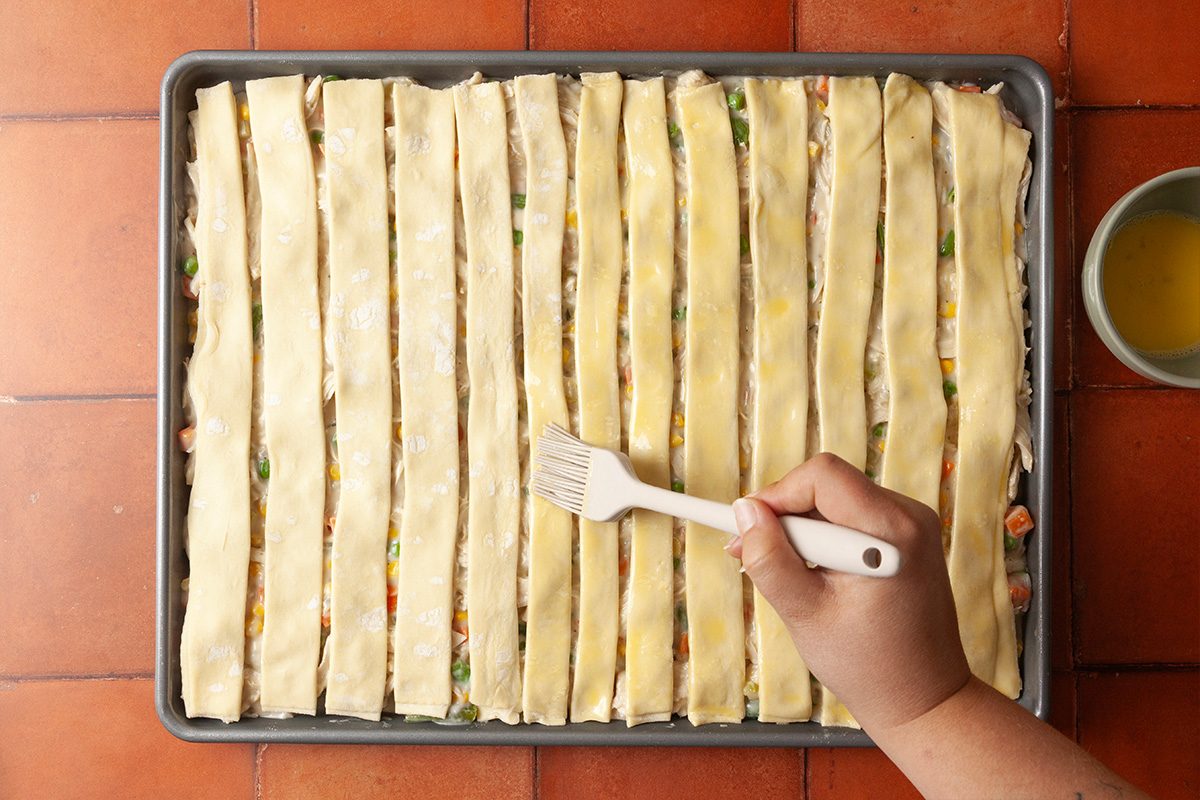 Overhead shot of a hand brushing egg wash onto strips of pastry dough laid over a rectangular baking dish filled with mixed vegetables on a tiled surface