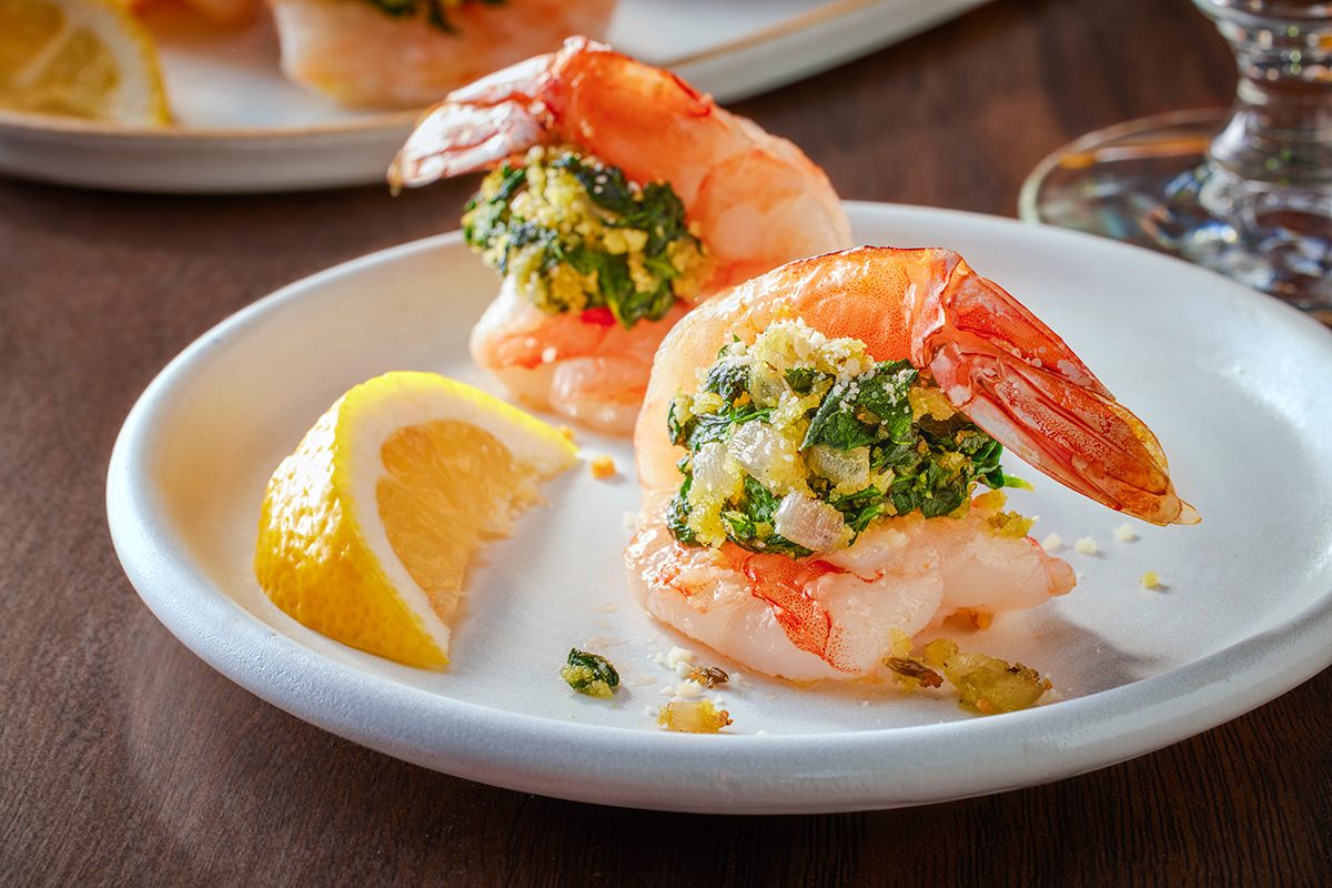 Two shrimp stuffed with a green and breadcrumb filling are served on a white plate with a lemon wedge. The shrimp are garnished and presented on a dark wooden table.
