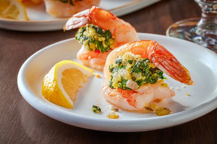 Two shrimp stuffed with a green and breadcrumb filling are served on a white plate with a lemon wedge. The shrimp are garnished and presented on a dark wooden table.