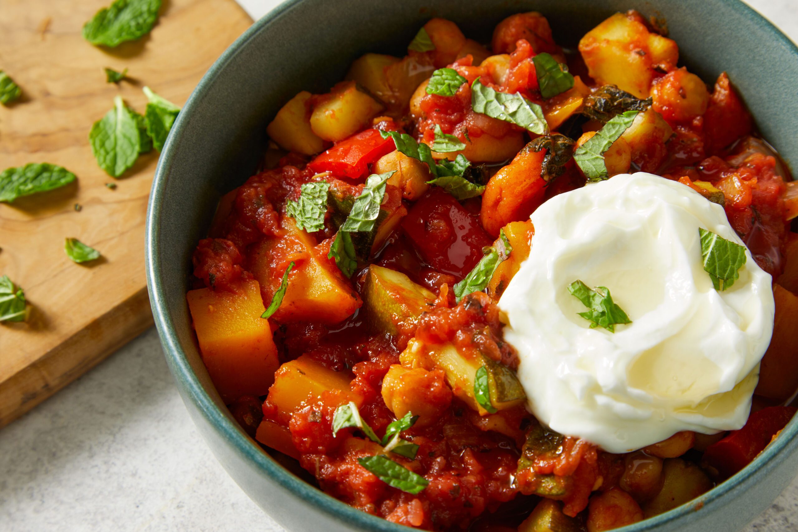A bowl of vegetable stew with chickpeas and tomatoes, topped with a dollop of yogurt and fresh chopped mint. The dish is served in a dark bowl on a light surface, with extra mint leaves on a wooden board nearby.
