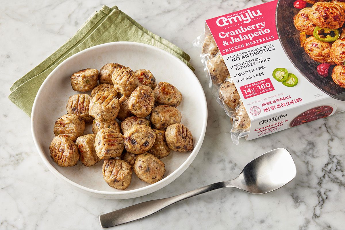 Overhead shot of a white bowl filled with grilled chicken meatballs on a marble surface; Beside it are a package of Amylu Cranberry & Jalapeño Chicken Meatballs, a folded green napkin, and a silver spoon