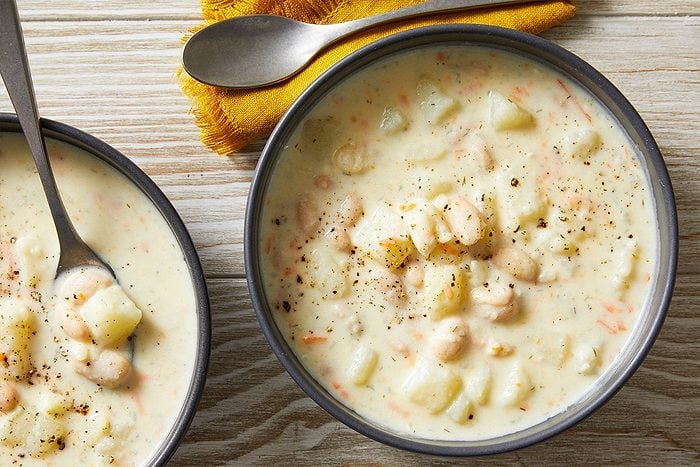 Overhead shot of two bowls of Potato Soup with Beans, featuring chunky potatoes and beans topped with black pepper, with spoons and a yellow napkin nearby;