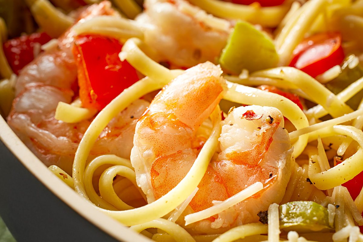 Close-up of cooked shrimp mixed with linguine pasta, diced red and green bell peppers, and sprinkled with herbs in a bowl.