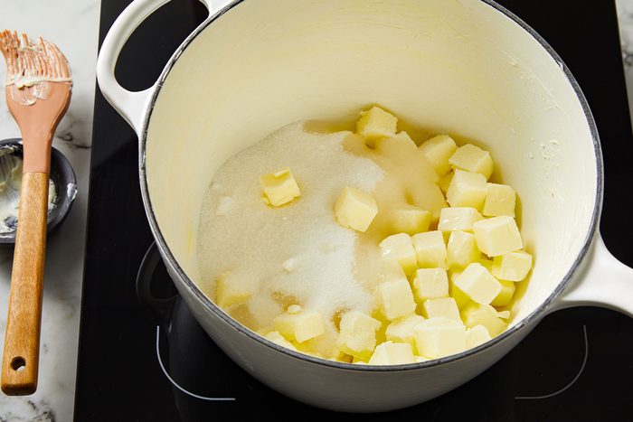 Overhead shot of butter cubes melting with sugar in a white saucepan on the stovetop, wooden spoon nearby.