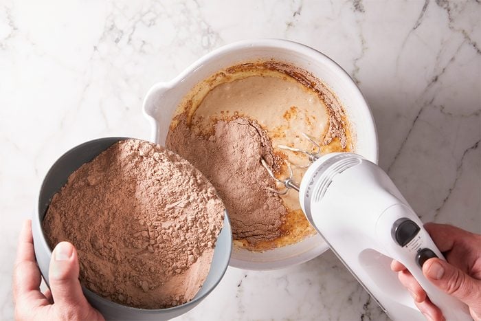 A person pours cocoa powder from a bowl into a mixing bowl with batter while using an electric hand mixer, on a white marble countertop.