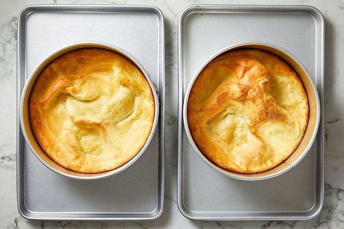 Overhead shot of two round, golden-brown Dutch baby pancakes in cake pans, side by side on a marble countertop, with puffed, uneven surfaces and lightly browned edges;