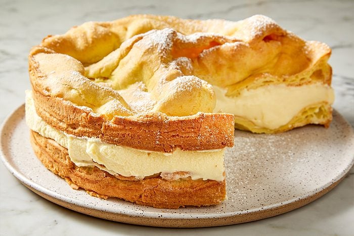 3/4 angle view shot of a slice of Karpatka Cake with creamy filling and a golden crust, dusted with powdered sugar, sitting on a plate on a marble surface;