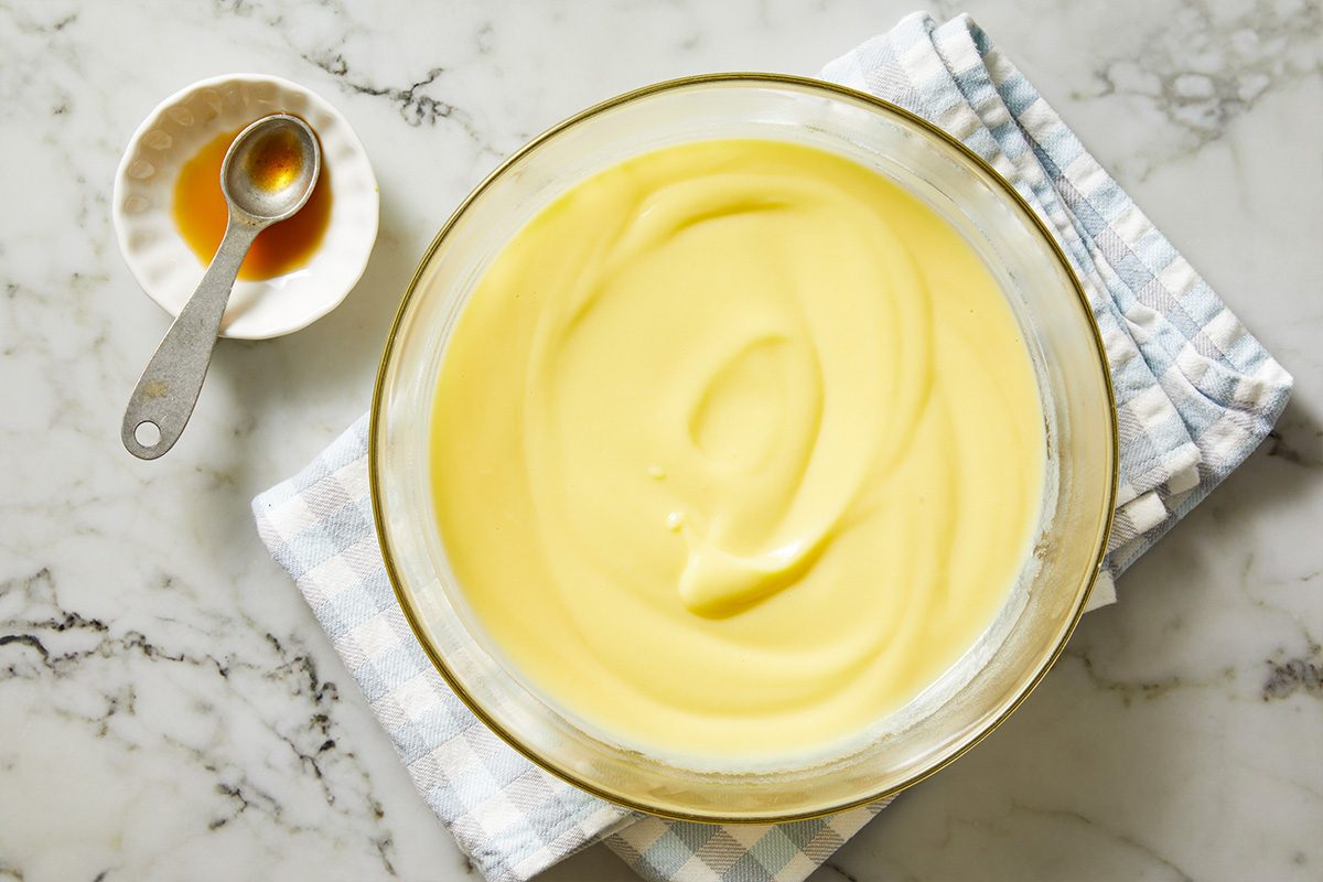 Overhead shot of a bowl of smooth, creamy yellow custard on a folded checkered cloth, with a small dish of vanilla extract and a measuring spoon beside it on a marble countertop;