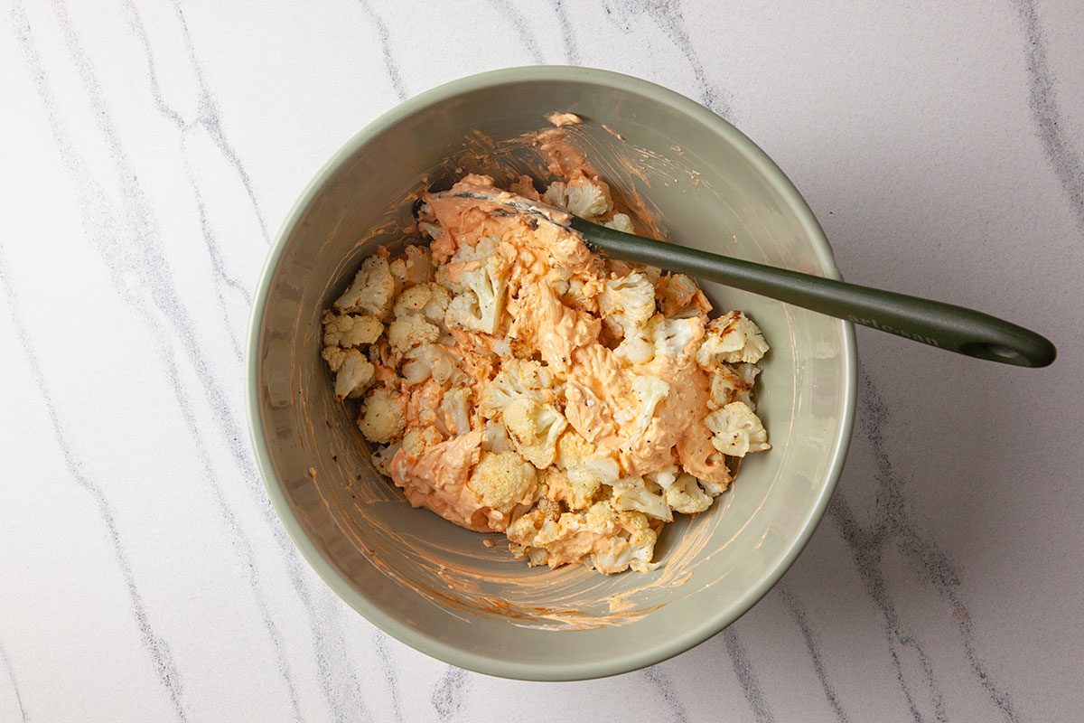 Overhead shot of roasted cauliflower florets tossed in a creamy orange sauce inside a bowl, with a spoon resting on a light marble surface;