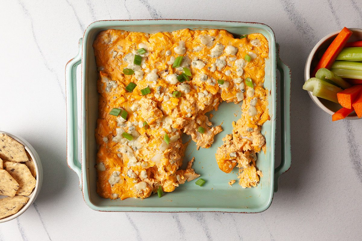 Overhead shot of buffalo cauliflower dip baked in a green ceramic dish, topped with melted cheese and green onions, with a scoop missing and veggie sticks on the side;