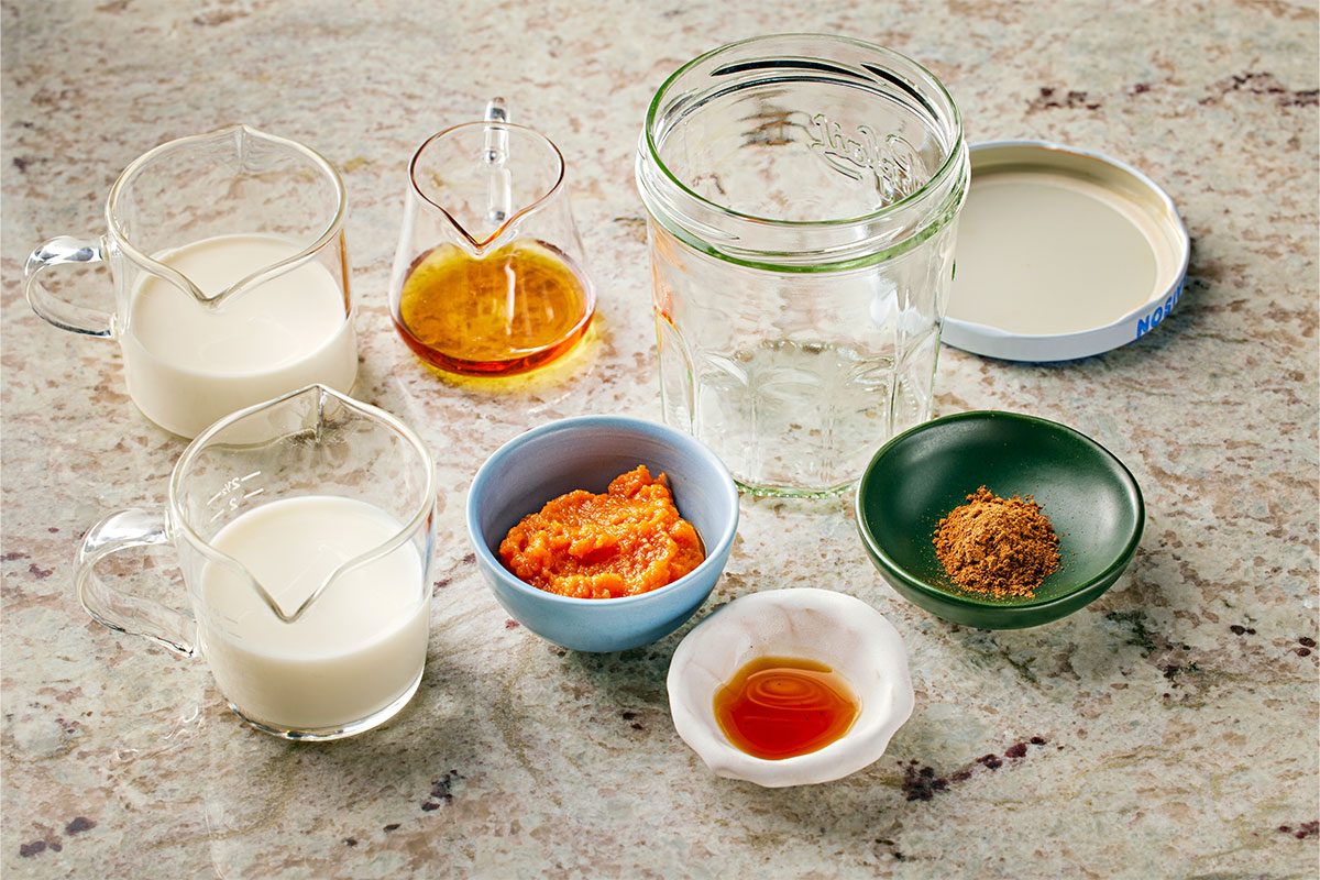 3/4 angle view shot of ingredients arranged on a counter, including two pitchers of milk, a glass jar with a lid, a small carafe of honey, a bowl of orange paste, a bowl of brown powder, and a dish of amber liquid;