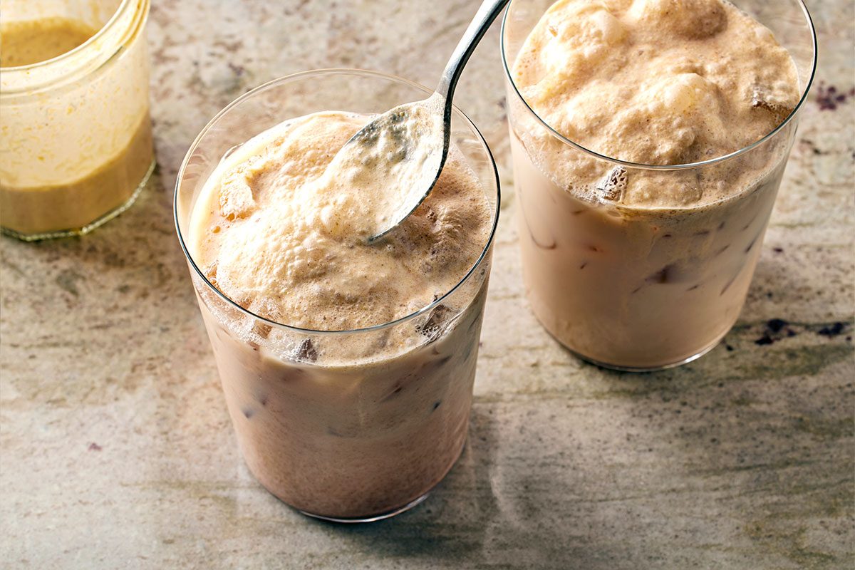 High angle shot of two glasses of iced coffee or milk-based drinks with frothy, creamy foam on top; one glass is being stirred with a spoon, both placed on a textured, light-colored surface;