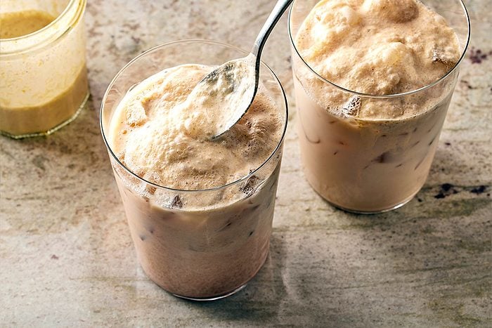 High angle shot of two glasses of iced coffee or milk-based drinks with frothy, creamy foam on top; one glass is being stirred with a spoon, both placed on a textured, light-colored surface;