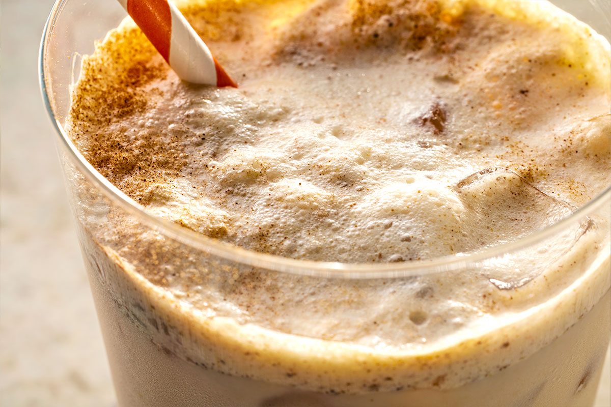 Closeup shot of a creamy, frothy Iced Pumpkin Cream Chai Tea Latte topped with cinnamon, served in a glass with a red-and-white straw;