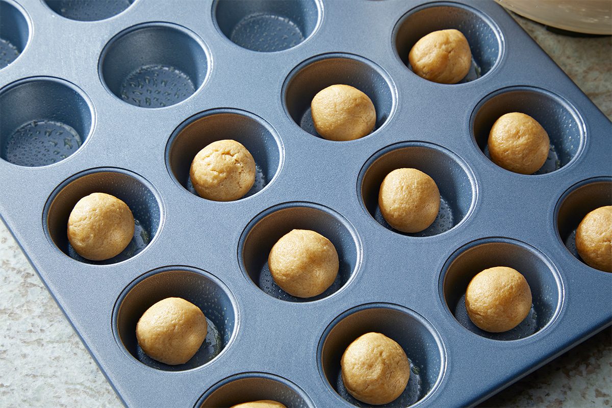 A metal mini muffin pan holds dough balls in each cup, ready for baking. The pan sits on a countertop.