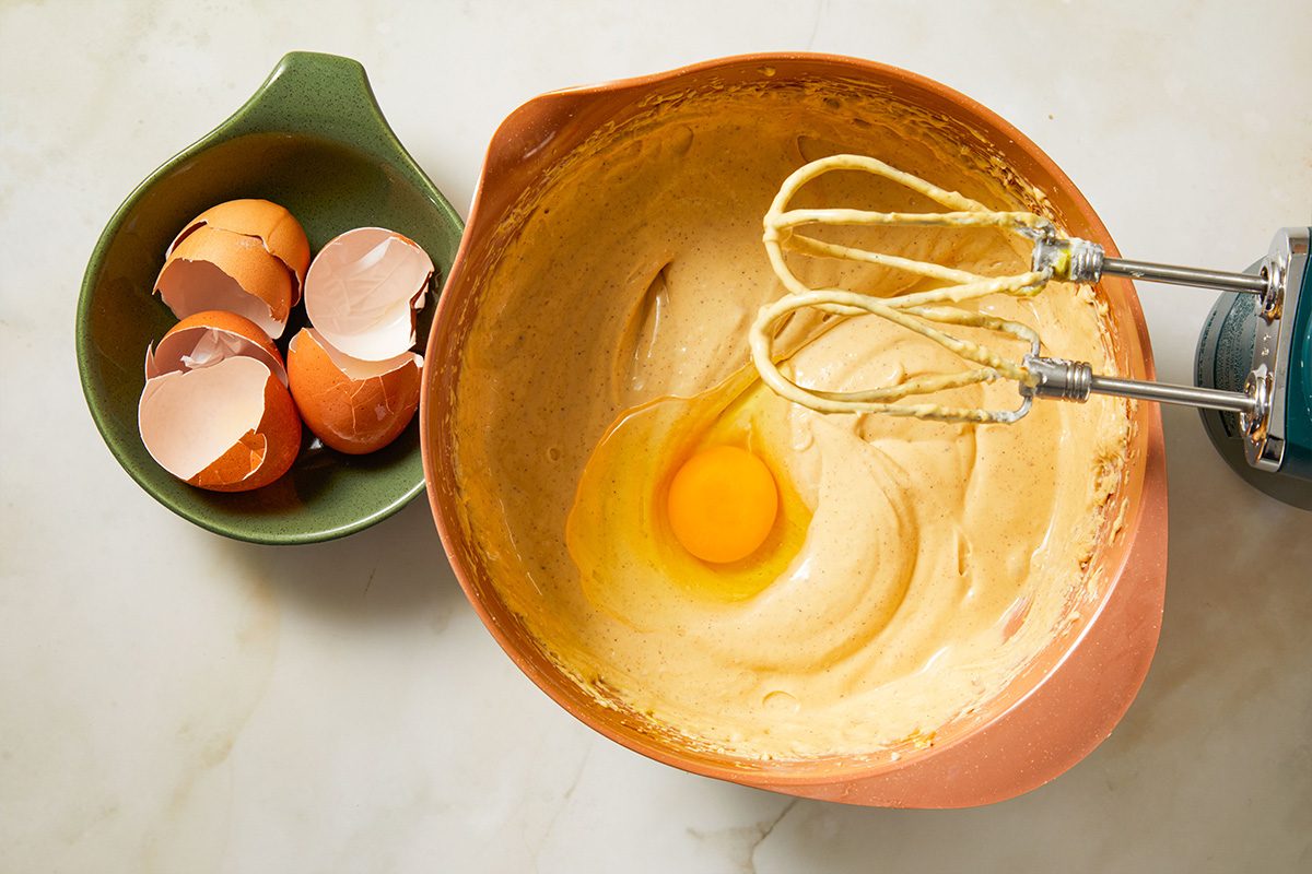 Overhead shot of eggs being mixed into the cheesecake batter, the mixture turning smooth and creamy under the electric beaters.
