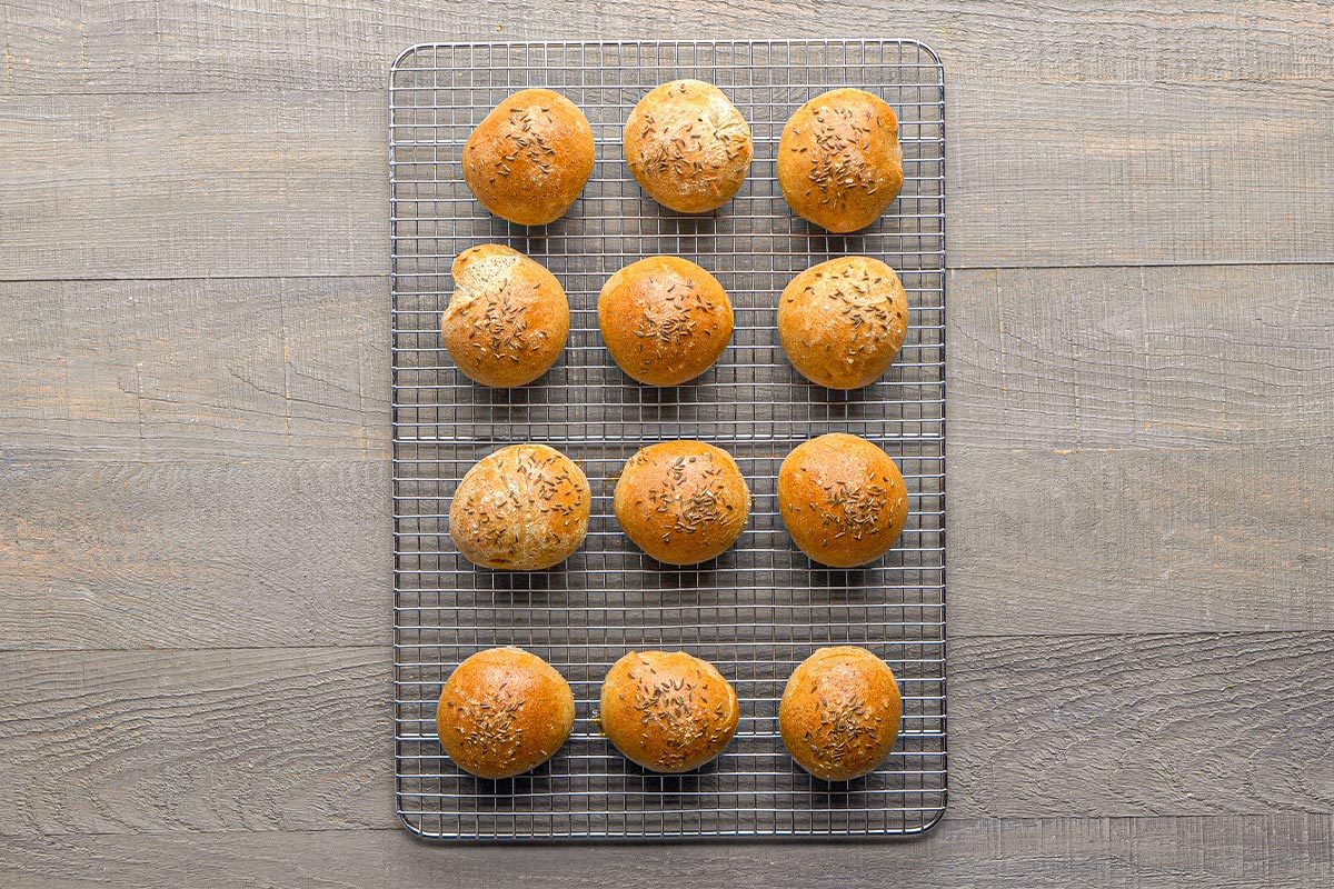 Overhead shot of twelve round bread rolls, some sprinkled with seeds, arranged in neat rows on a wire cooling rack on a wooden surface. One roll has a bite taken out of it;