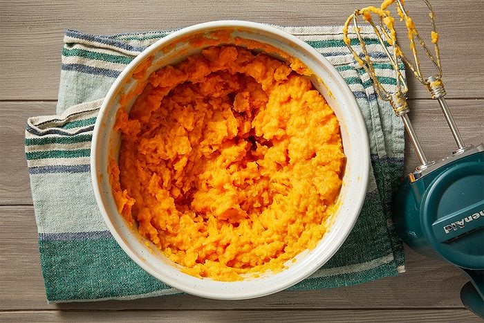 Overhead shot of a bowl of mashed sweet potatoes on a striped kitchen towel, with an electric hand mixer resting beside it on a wooden surface;