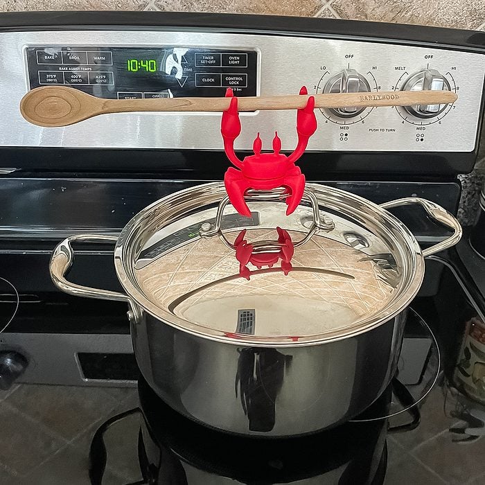 A red crab-shaped utensil holder is clipped to the rim of a stainless steel pot, holding a wooden spoon horizontally above the pot