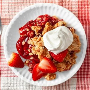 A plate of strawberry crumble topped with whipped cream, garnished with fresh strawberry slices, sits on a white dish atop a red and white checkered tablecloth.