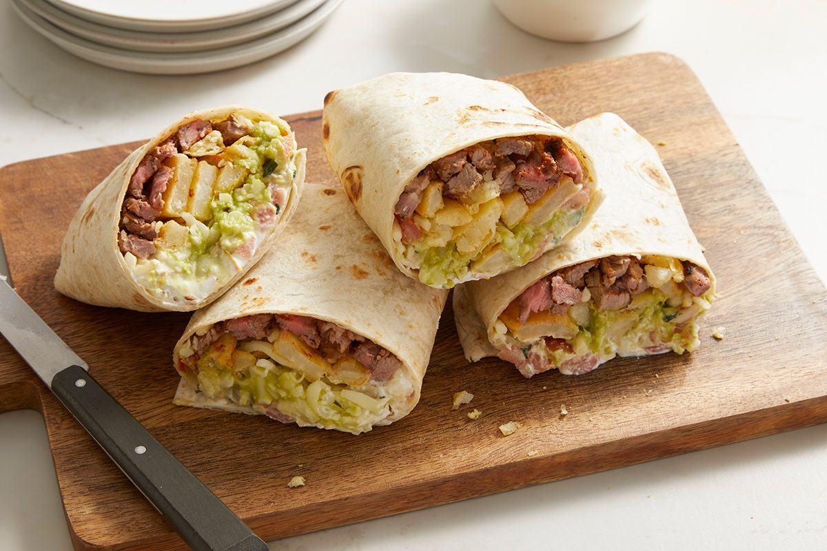 Four burritos cut in half reveal fillings of meat, lettuce, fries, and cheese, arranged on a wooden cutting board with a knife beside them. White plates are stacked in the background.