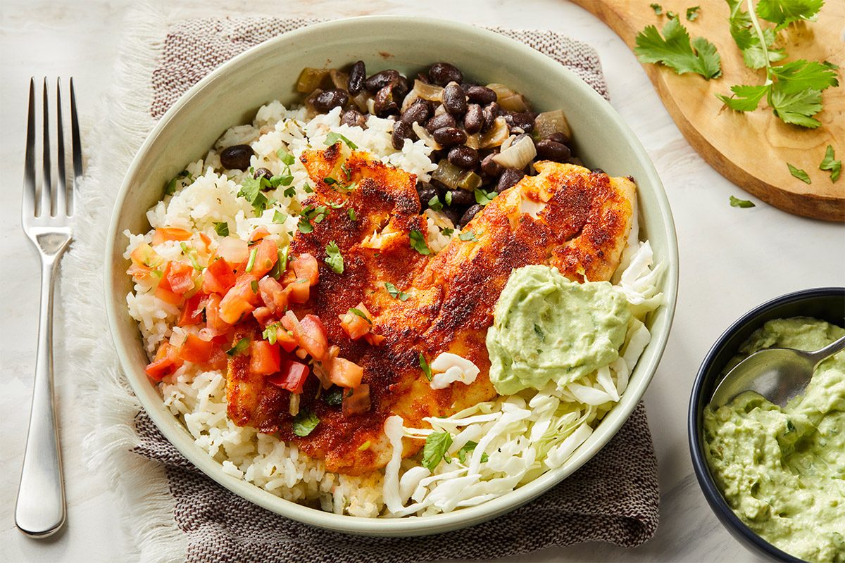 Plated fish taco bowl with tilapia, rice, beans, cabbage, pico de gallo, and avocado crema, styled for serving with a spoon and napkin.
