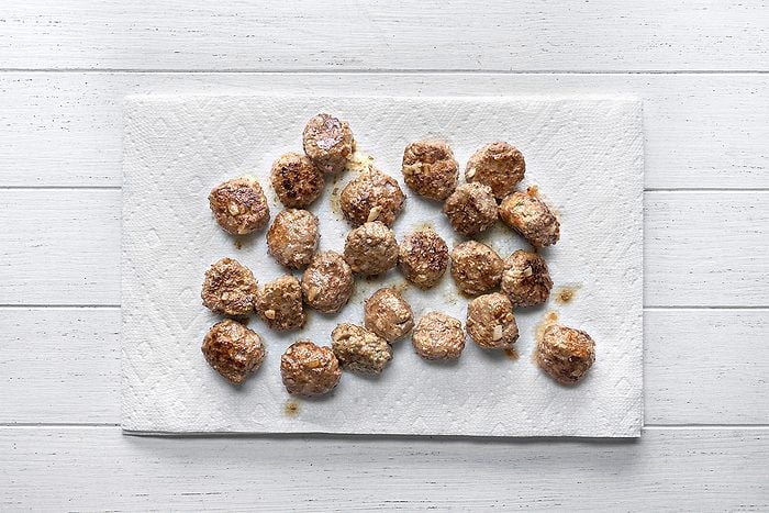 overhead shot of Several cooked meatballs are arranged on a white paper towel, which is placed on a white wooden surface, The meatballs appear browned and slightly oily