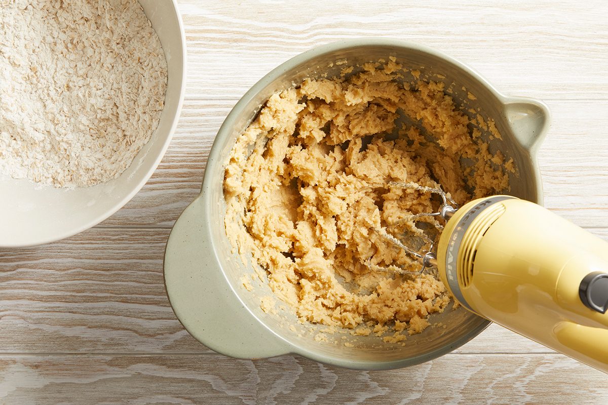 A yellow hand mixer blends cookie dough in a mixing bowl, with a separate bowl of dry ingredients visible beside it on a light wood surface.