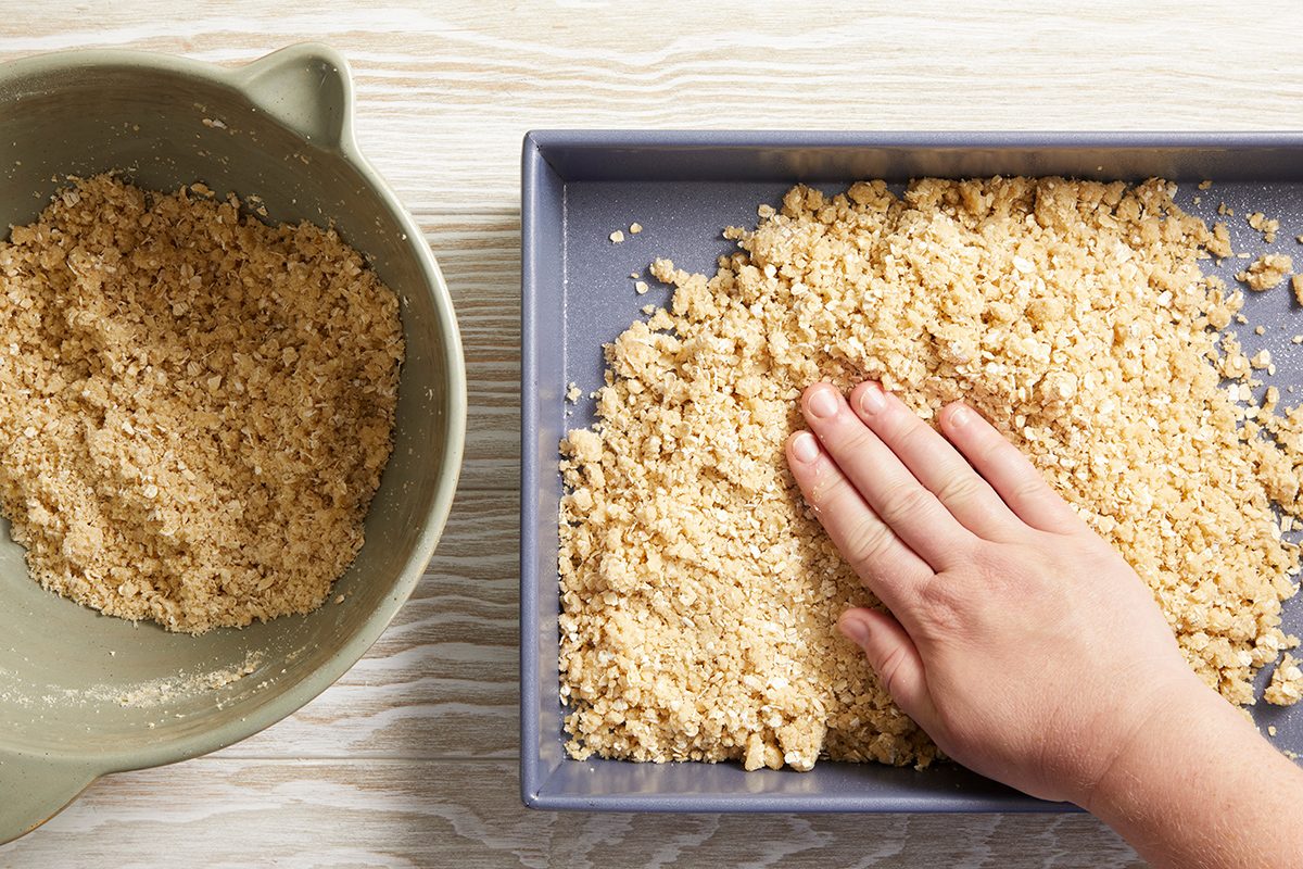 A hand presses crumbly dough mixture into a square baking pan, with extra dough in a green mixing bowl beside it on a light-colored surface.