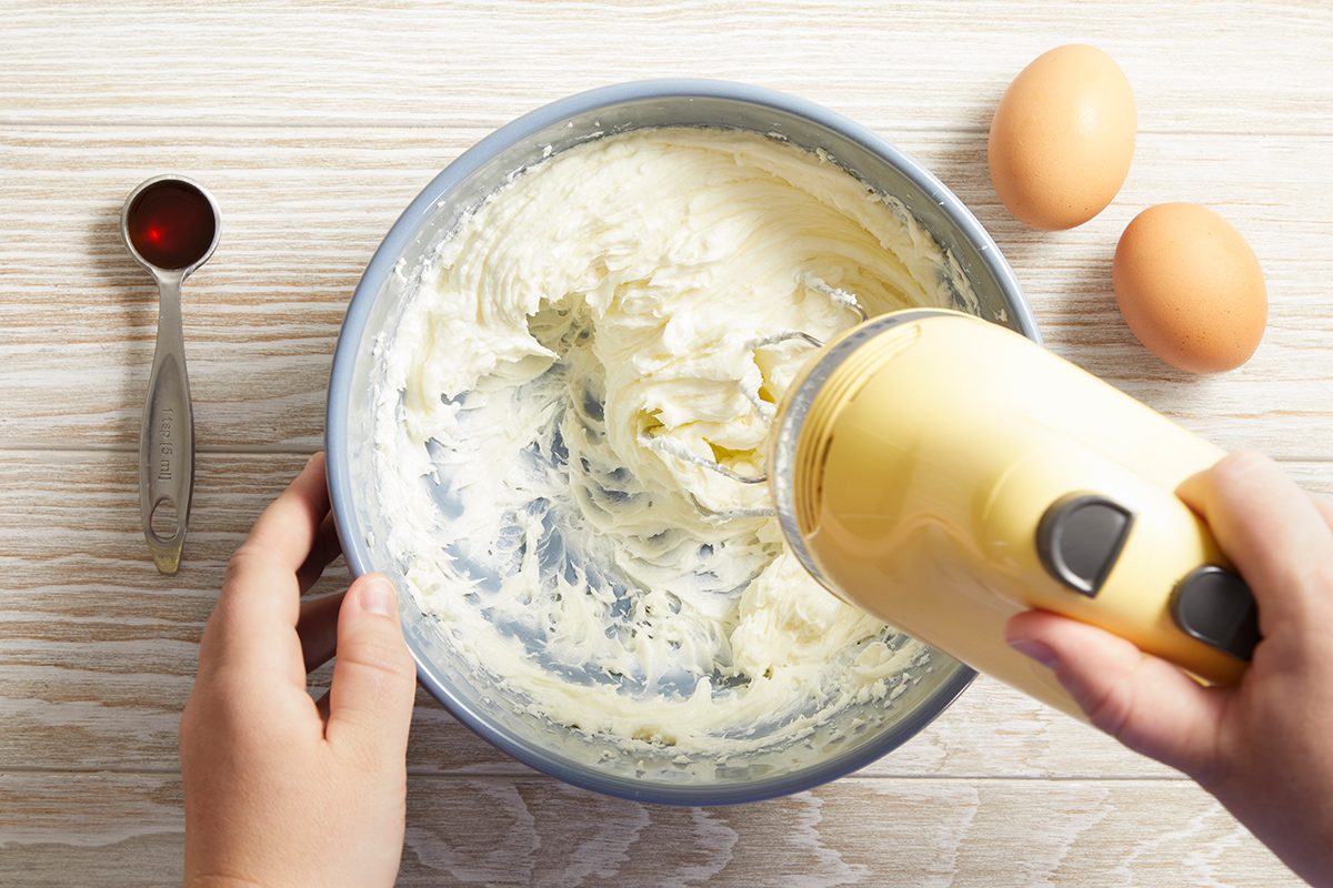 A person uses a yellow hand mixer to blend cream in a mixing bowl. Nearby are two eggs and a measuring spoon with red liquid, all on a light wooden surface.