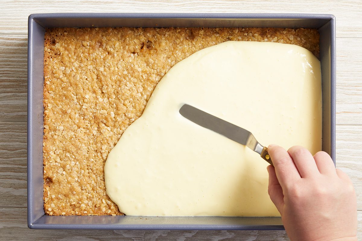 A hand uses a spatula to spread creamy white frosting over a baked oat crust in a rectangular metal pan on a light wooden surface.