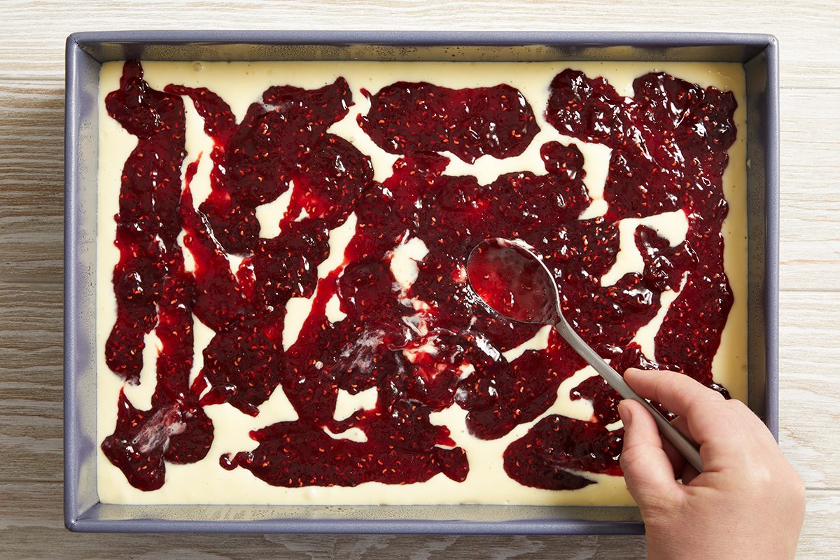 A hand uses a spoon to spread a layer of red berry jam over a light-colored batter in a rectangular baking pan on a light wooden surface.
