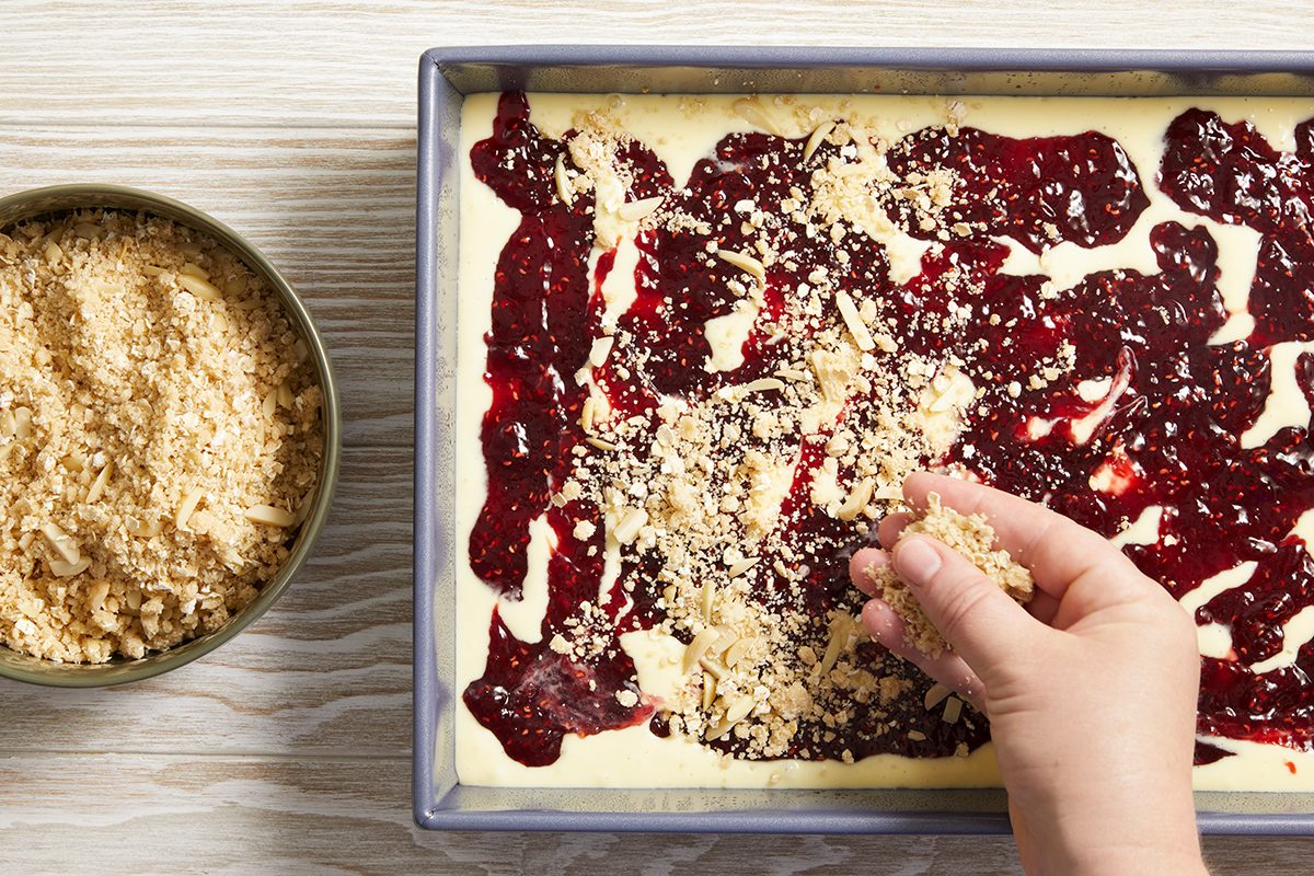 A hand sprinkles crumb topping over a rectangular pan filled with cake batter and swirls of red fruit jam. A bowl of additional crumb topping sits beside the pan on a light wooden surface.