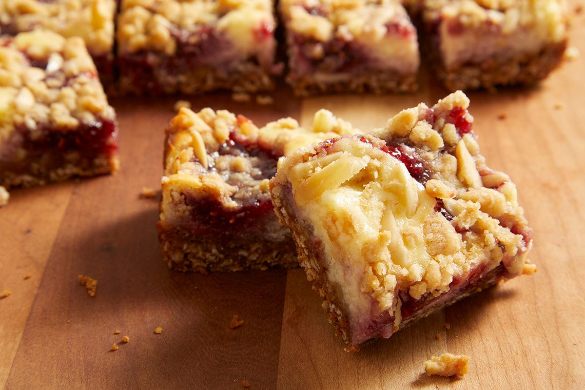 Several square dessert bars with a crumbly oat topping and a visible layer of red fruit jam are arranged on a wooden surface, with one bar slightly tilted to show its layers.