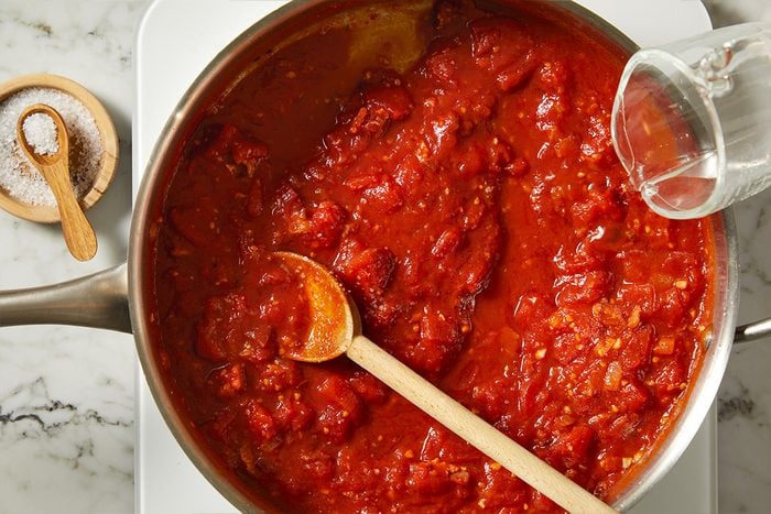Overhead shot of crushed whole plum tomatoes added to the skillet, forming a vibrant red sauce as vodka is poured in. The tomato mixture simmers with bacon and aromatics, beginning to transform into a classic vodka sauce.