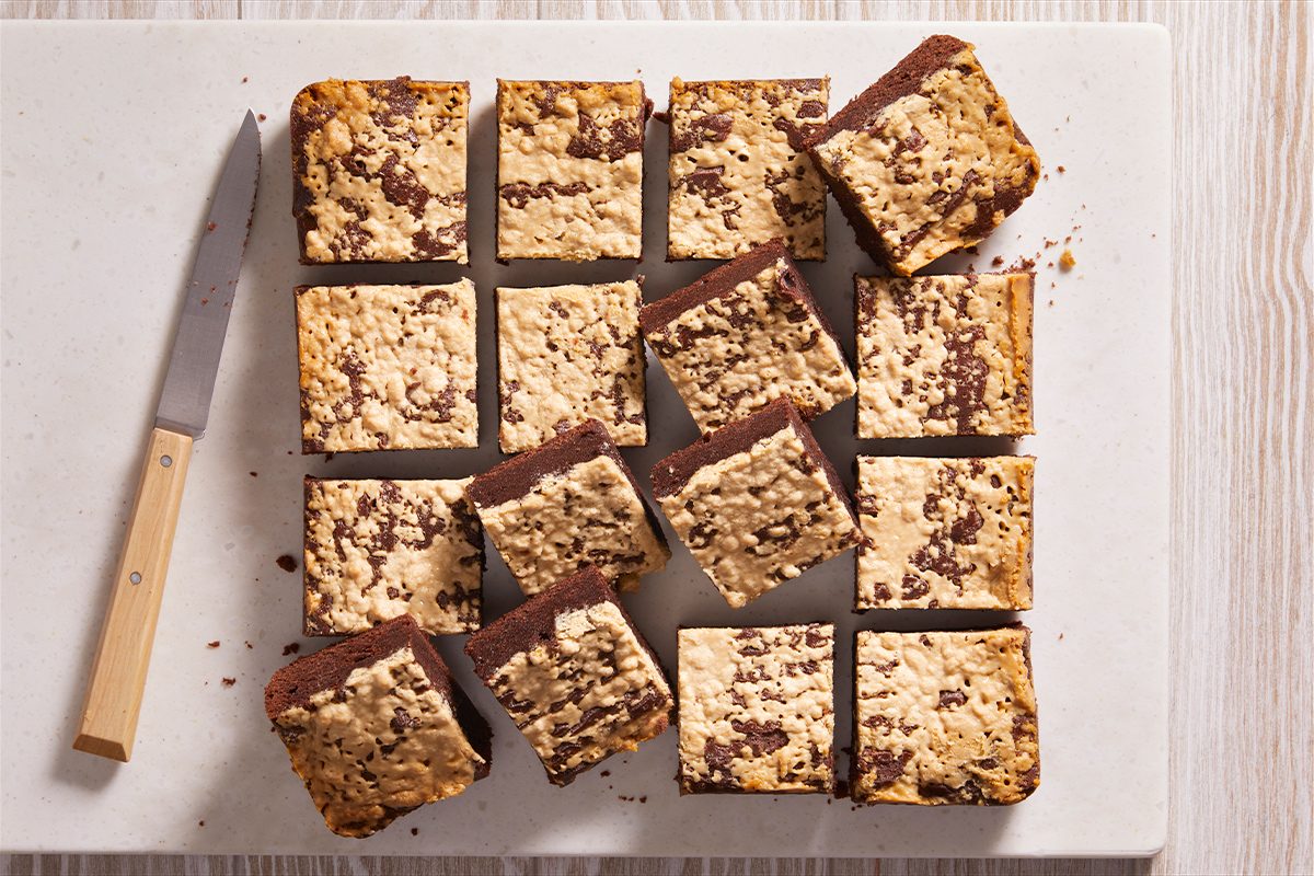 Fifteen chocolate oat bars are arranged on a white cutting board next to a knife with a wooden handle. The bars have a crumbly oat topping and are cut into neat squares.