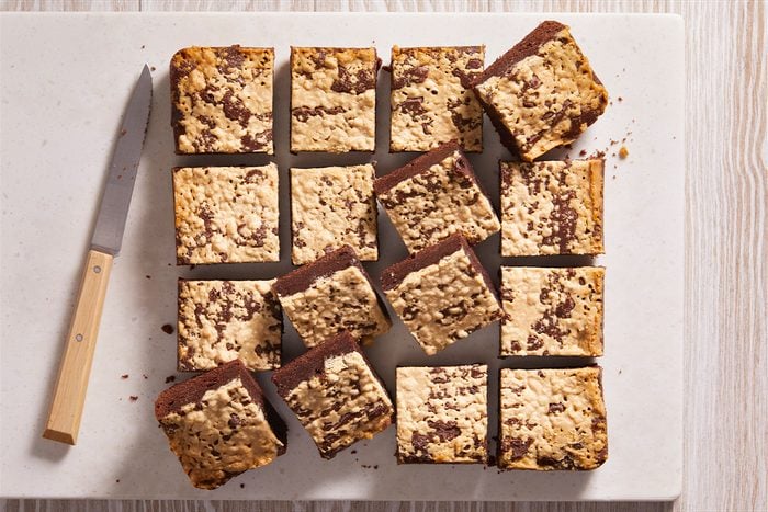 Fifteen chocolate oat bars are arranged on a white cutting board next to a knife with a wooden handle. The bars have a crumbly oat topping and are cut into neat squares.