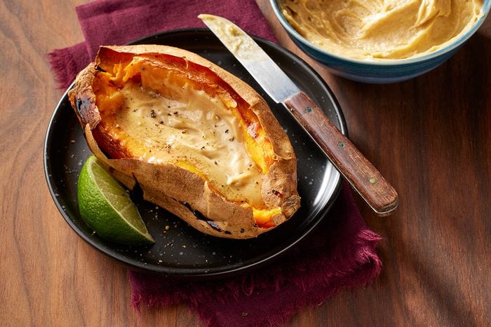 A baked sweet potato split open and filled with melted butter sits on a black plate with a lime wedge, next to a knife and a bowl of butter. The plate is on a wooden table with a burgundy napkin underneath.