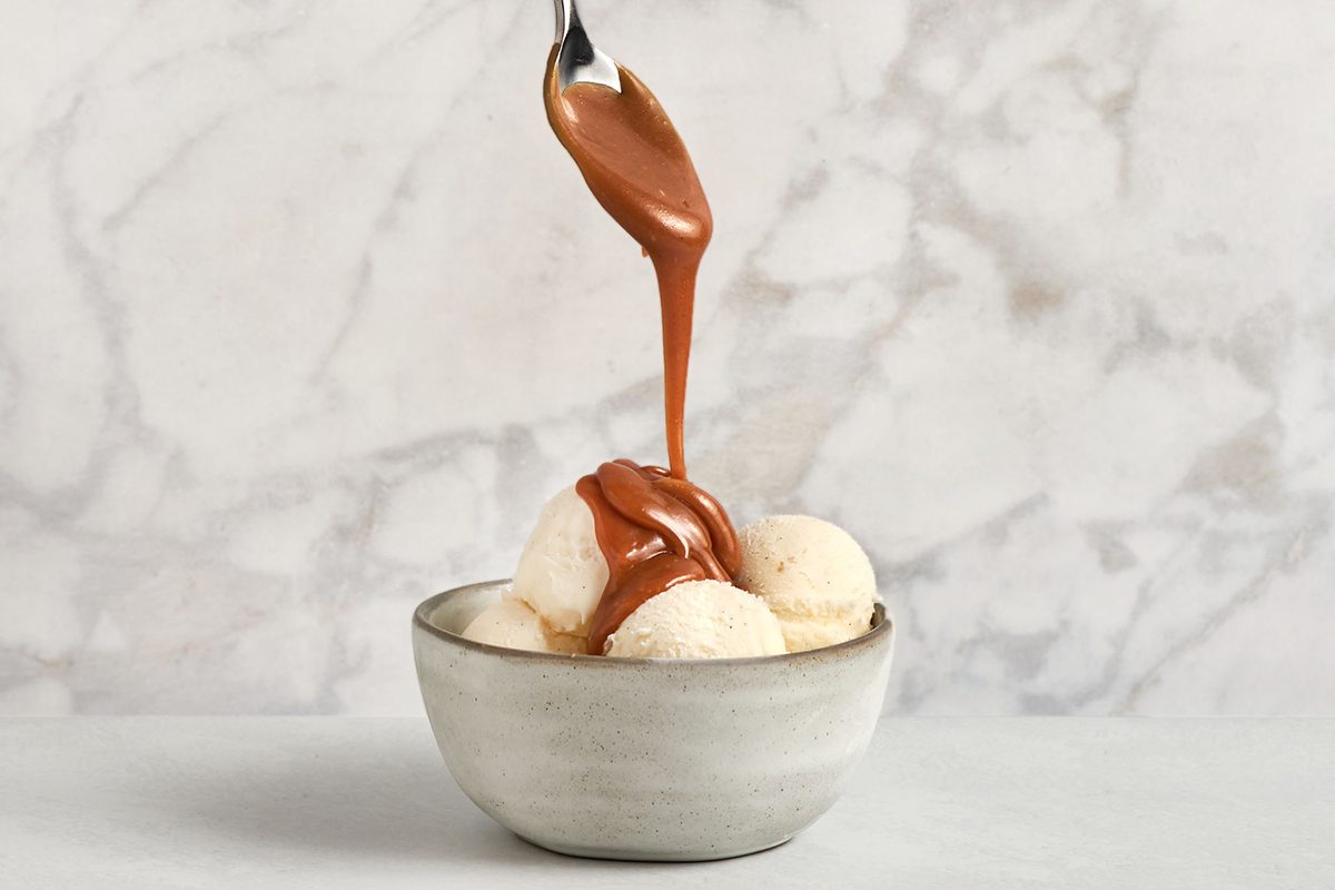 A spoon drizzles caramel sauce over three scoops of vanilla ice cream in a light gray bowl, set against a marble background.