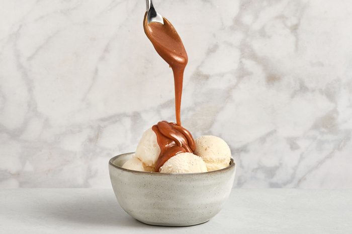 A spoon drizzles caramel sauce over three scoops of vanilla ice cream in a light gray bowl, set against a marble background.