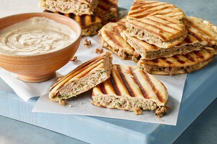 Grilled sandwiches filled with ground meat and herbs are stacked on parchment paper next to a bowl of creamy dipping sauce on a light blue cutting board.
