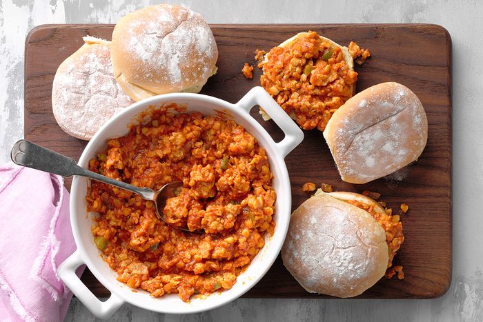 A bowl of sloppy joe filling with a spoon sits on a wooden board, surrounded by four sandwich buns, some filled with the mixture and some plain. A pink cloth is partially visible to the side.