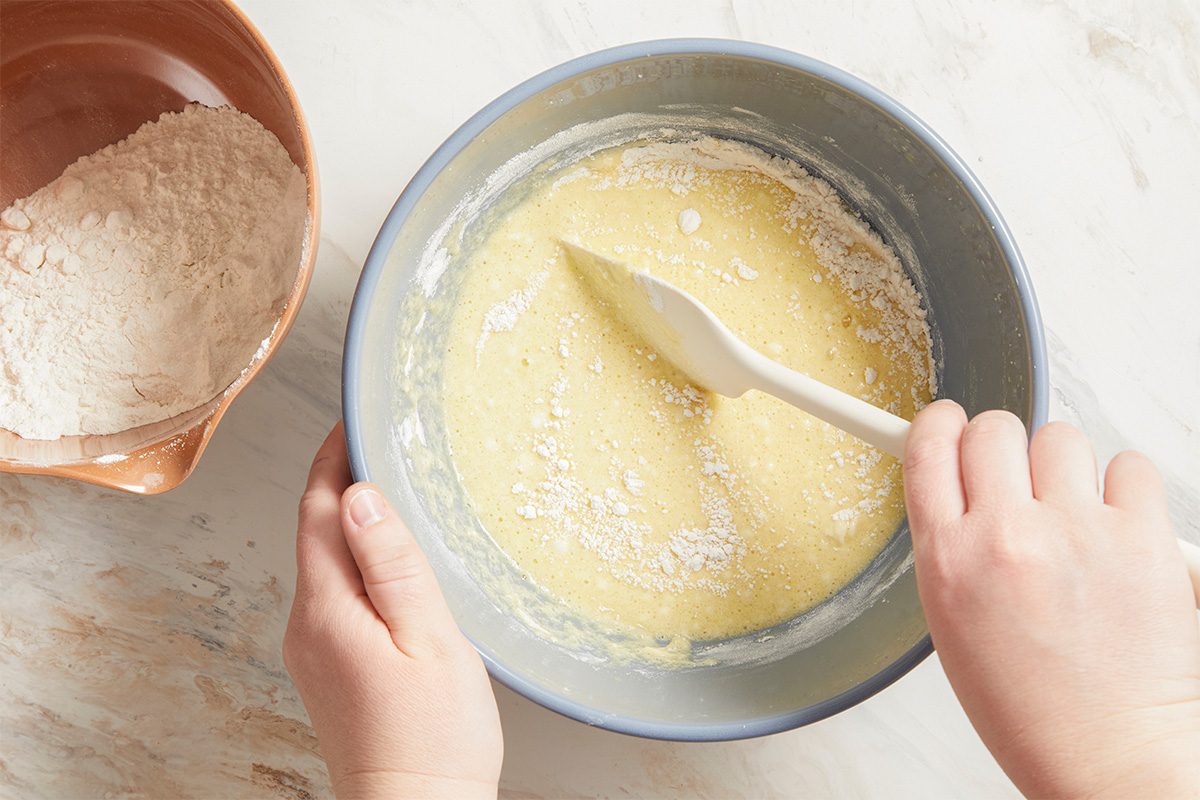 Overhead action shot of hands stirring apricot bread batter in a mixing bowl. The softened butter, sugar, egg, and orange juice are being combined into a smooth mixture on a light countertop.