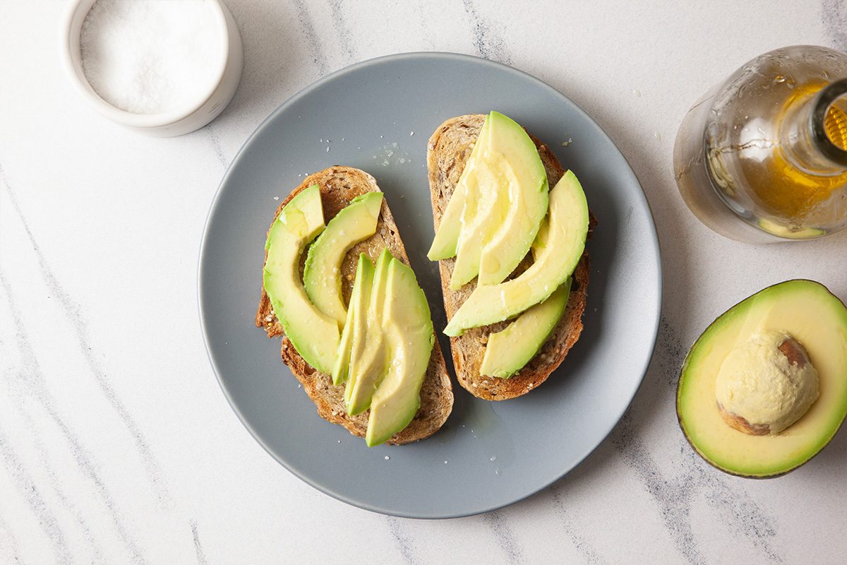 Overhead horizontal image of avocado toast with evenly layered avocado slices covering toasted bread. Supporting ingredients are styled around the plate to highlight a clean, balanced presentation.