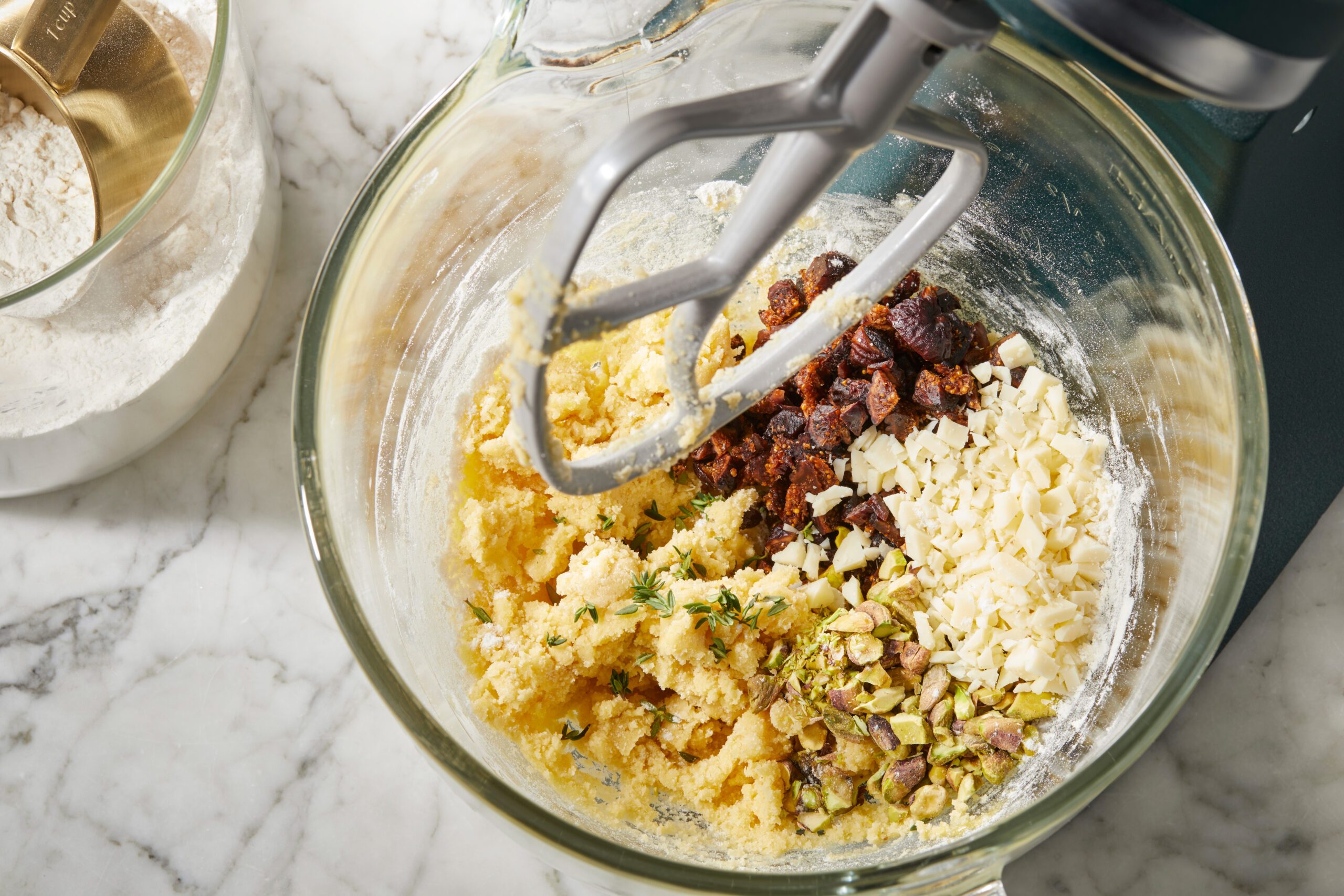 Top-down image of biscuit dough being mixed with chopped dried figs, pistachios, Pecorino Romano Parmesan cheese, and fresh thyme in a stand mixer bowl.