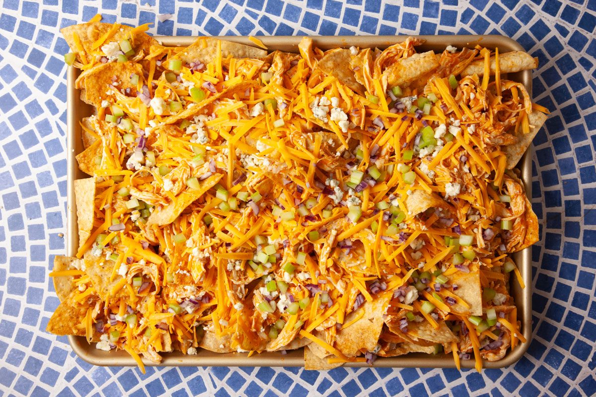 Overhead image of Buffalo Chicken Nachos topped with shredded cheddar, blue cheese crumbles, chopped celery, and red onion. This stage captures the addition of fresh vegetables before finishing with dressing drizzle.